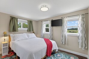 Ein Schlafzimmer mit Bett, Nachttisch, Lampe, Fernseher und Fenstern mit Vorhängen.