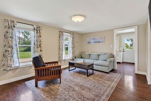 A living room with a wooden chair, a sofa, a coffee table, and a rug. There are two windows with curtains, a door, and a painting on the wall.