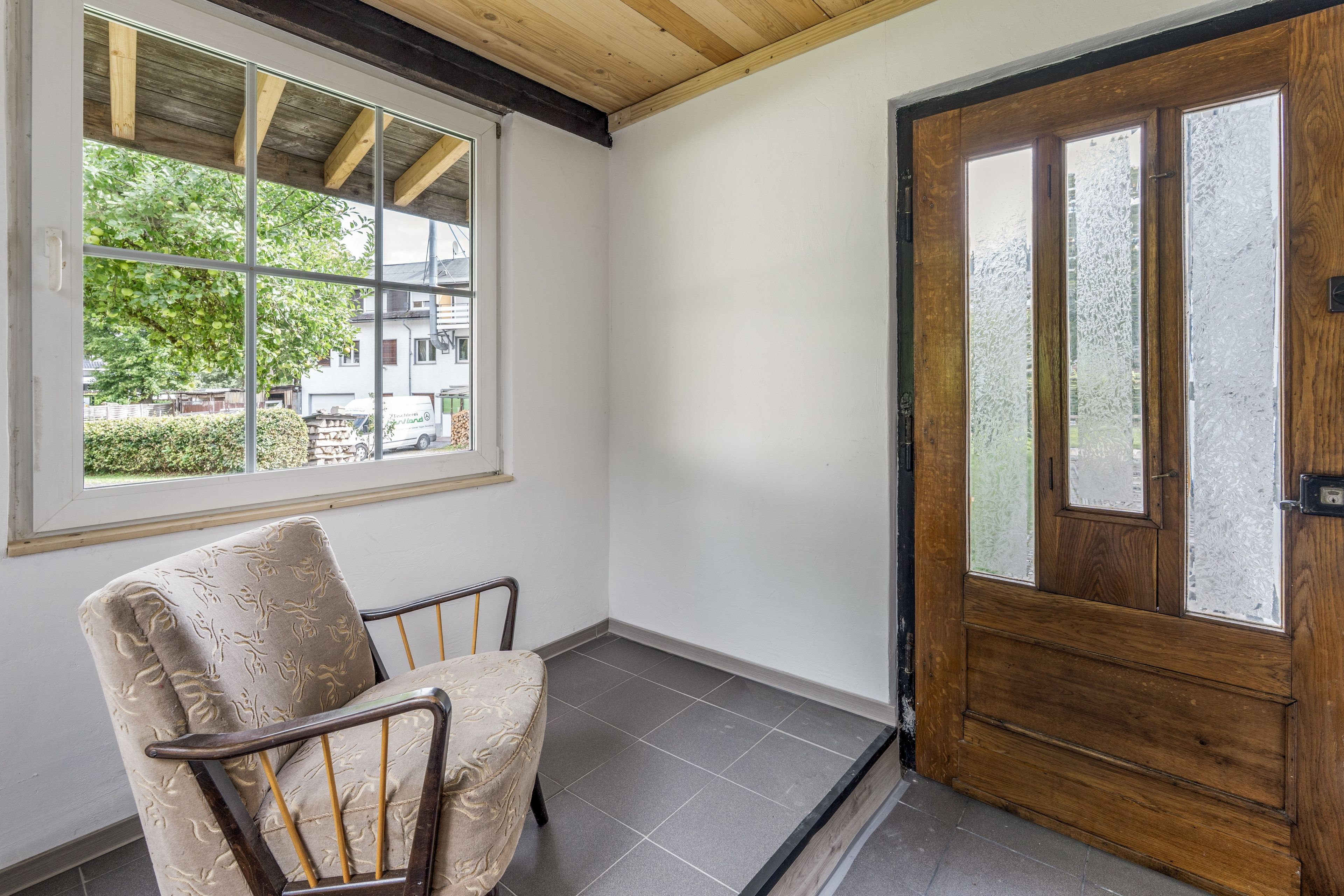 Una habitación con puerta de madera, ventana con vista a follaje y una silla.