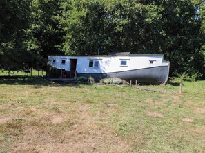 A stationary boat with a small attached structure, possibly a cabin, in a grassy area with trees in the background. - Ladies of Bolton (Norwich)
