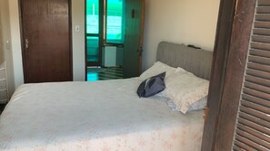 Ein Bett mit gemustertem Überwurf und Kissen, eine Holztür und Blick auf einen Raum mit Fliesenboden und einem Fenster.