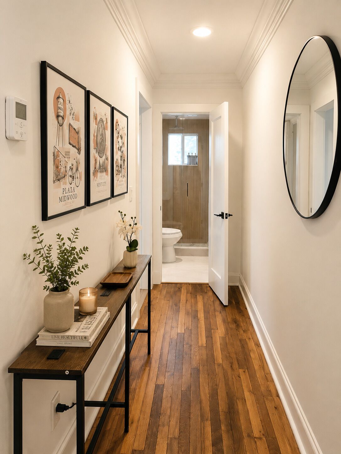 A hallway with wooden flooring, a mirror, framed pictures on the wall, a small table with a plant, and a door leading to a bathroom.