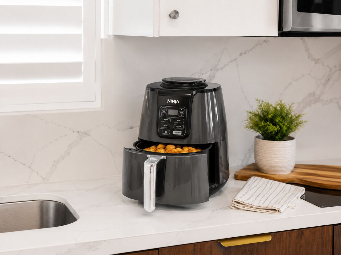 Une cuisine équipée d’un appareil de cuisson à air chaud Ninja noir, d’un plan de travail en marbre, d’un évier en acier inoxydable, d’une planche à découper en bois et d’une plante en pot.