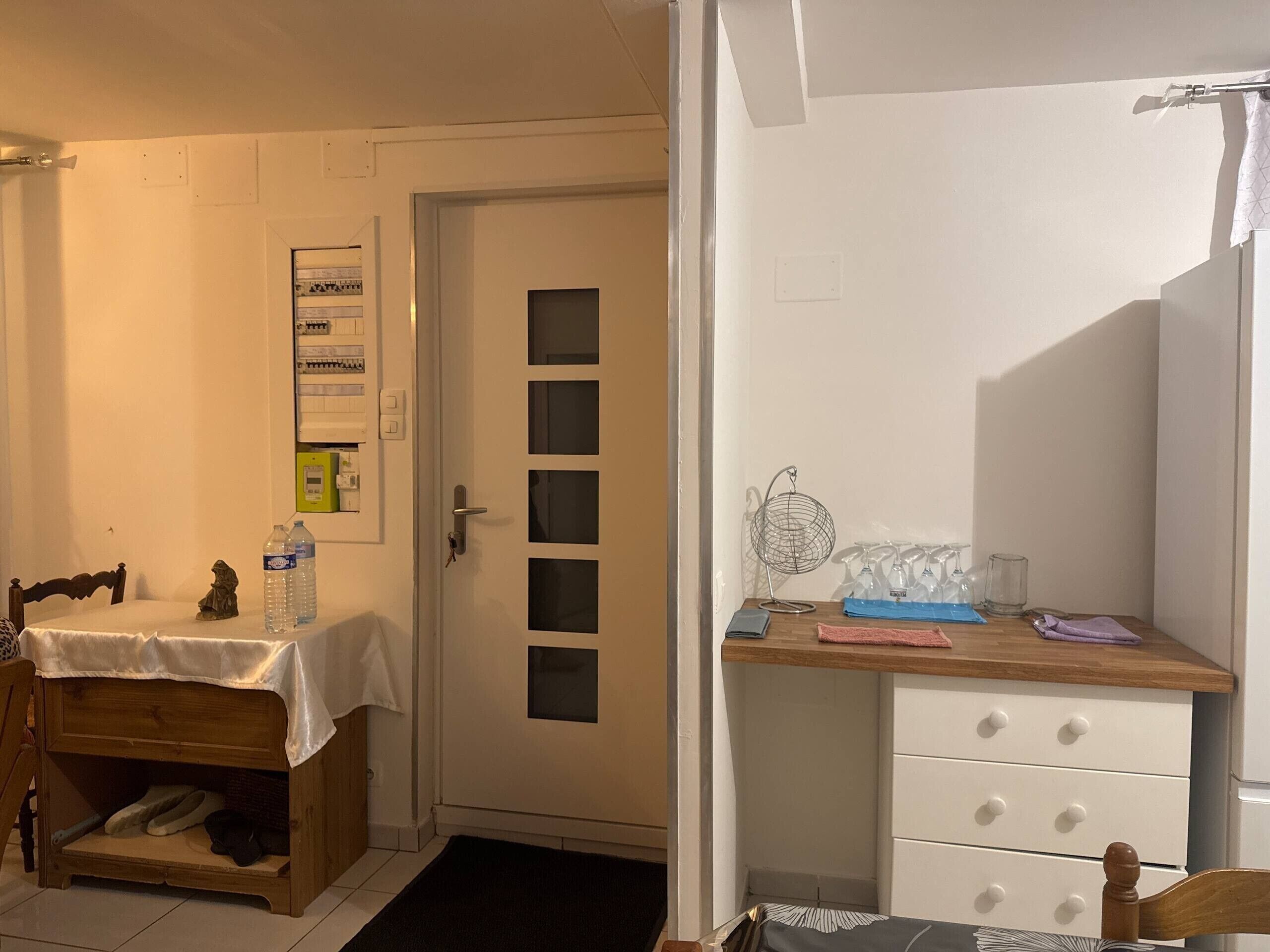 A room with a wooden desk, a chair, a bed with a white headboard, a door with a window, and a small shelf with items.