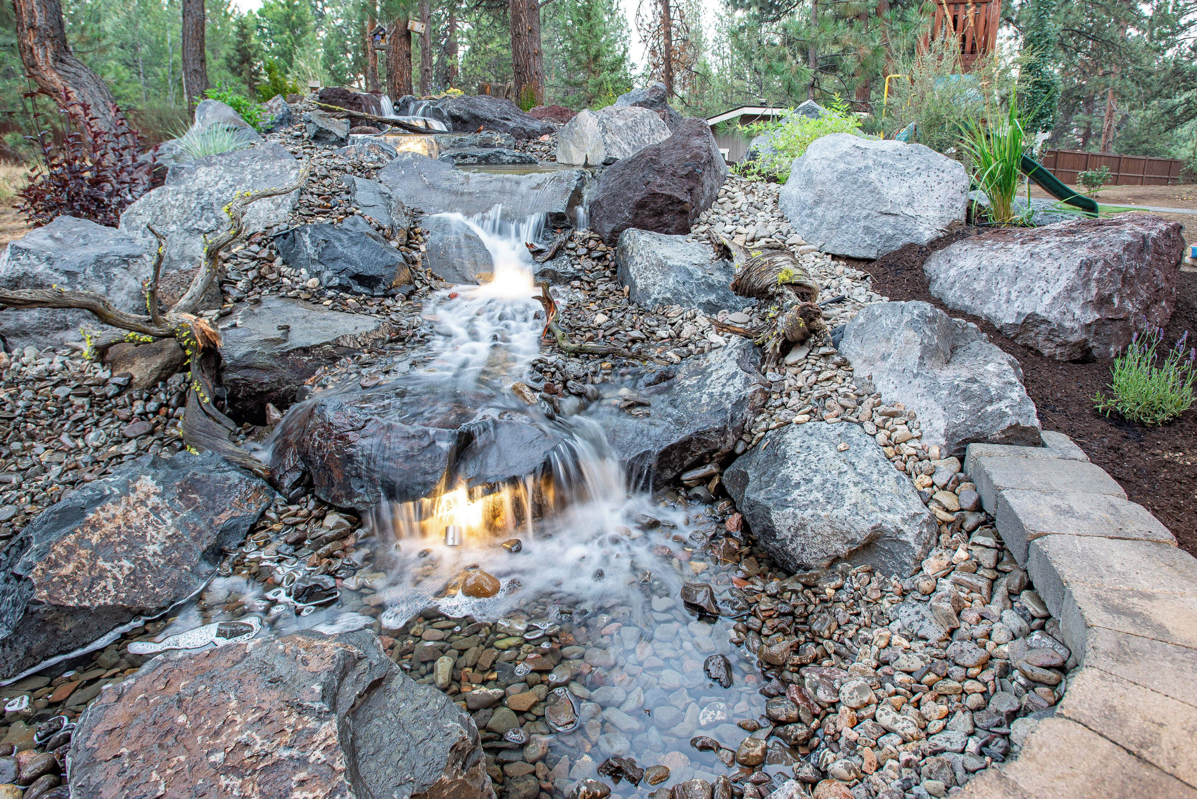 Una característica acuática natural con un arroyo en cascada sobre rocas, rodeada de un jardín ajardinado con diversas plantas y una terraza de madera al fondo.