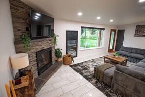 Ein Wohnzimmer mit Kamin, großem Flachbildfernseher, einer Couch und Blick ins Freie.
