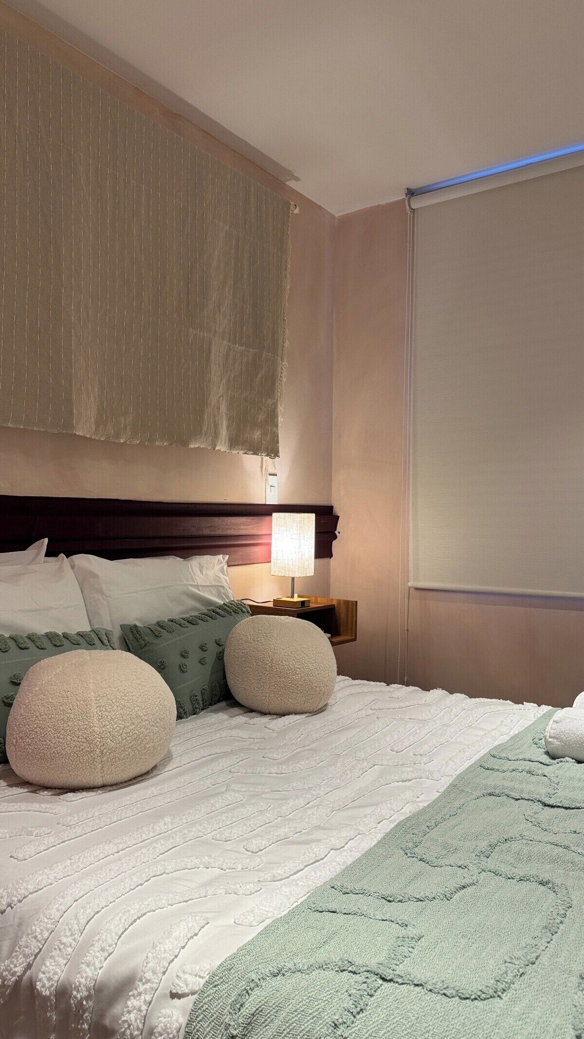 Une chambre d’hôtel avec un lit, des oreillers, une table de chevet, une lampe et une fenêtre avec des stores.