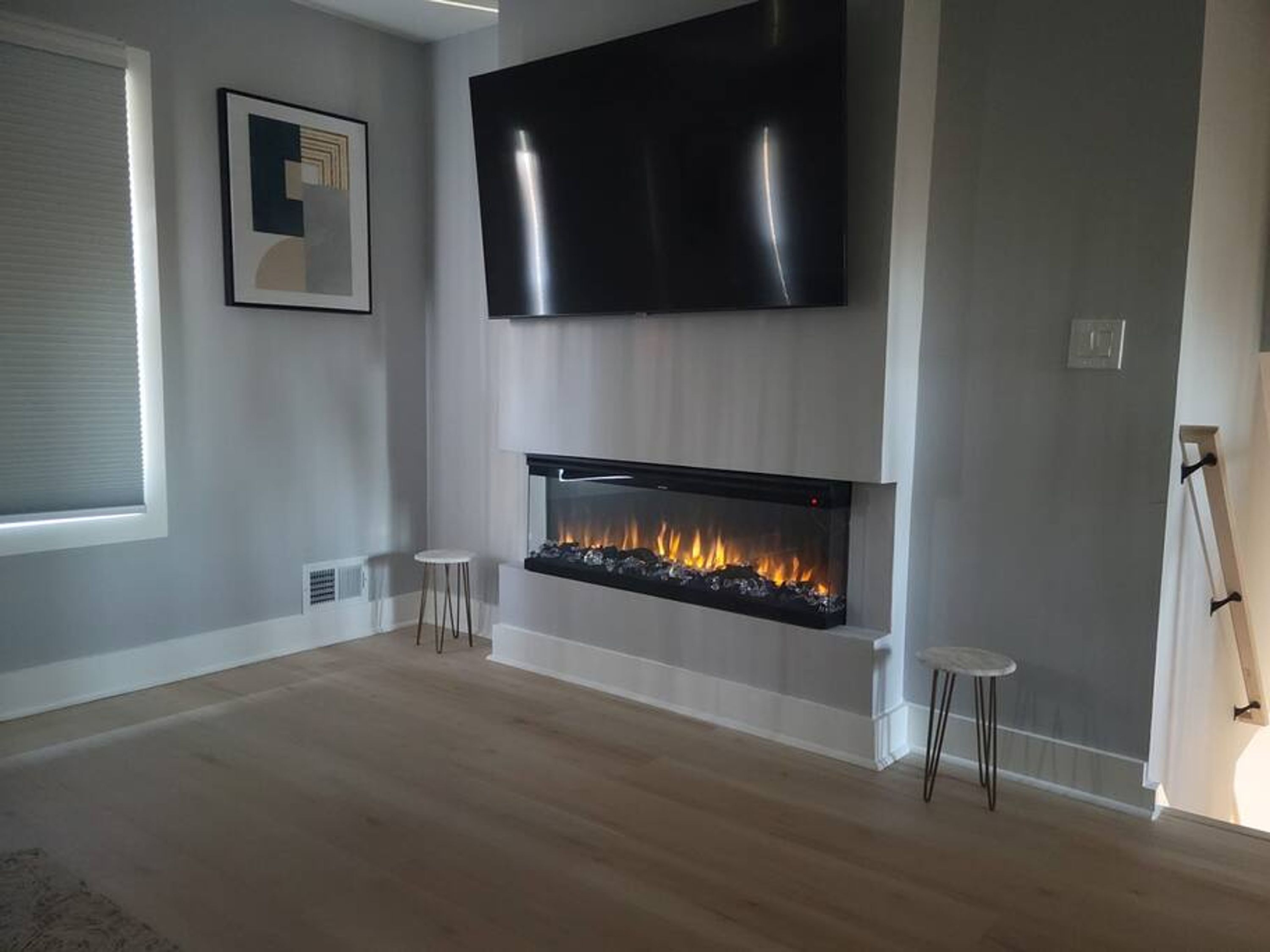 A modern living room with a flat-screen TV mounted above a fireplace, two small stools, and a framed picture on the wall.
