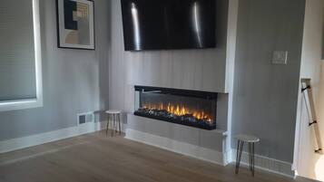 A modern living room with a flat-screen TV mounted above a fireplace, two small stools, and a framed picture on the wall.