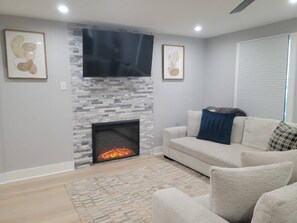 A living room with a fireplace, a flat-screen TV mounted above it, a light-colored sofa with cushions, and a patterned rug on the floor. - Urban Xcape- Family Vibe With Plush Amenities (Silver Spring)