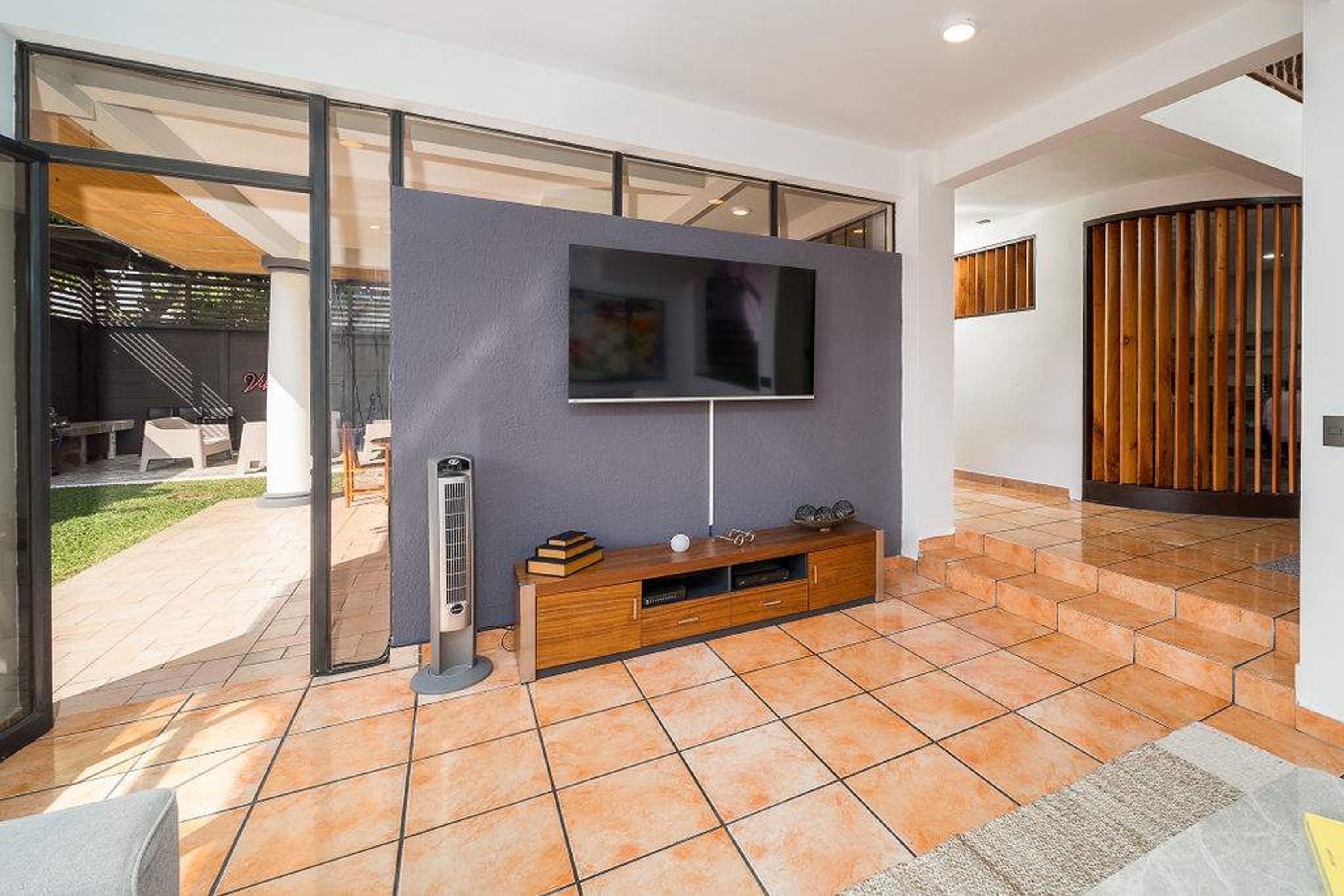 A modern living room with a flat-screen TV mounted on a wall, a wooden entertainment unit below, and a large glass door leading to an outdoor area.