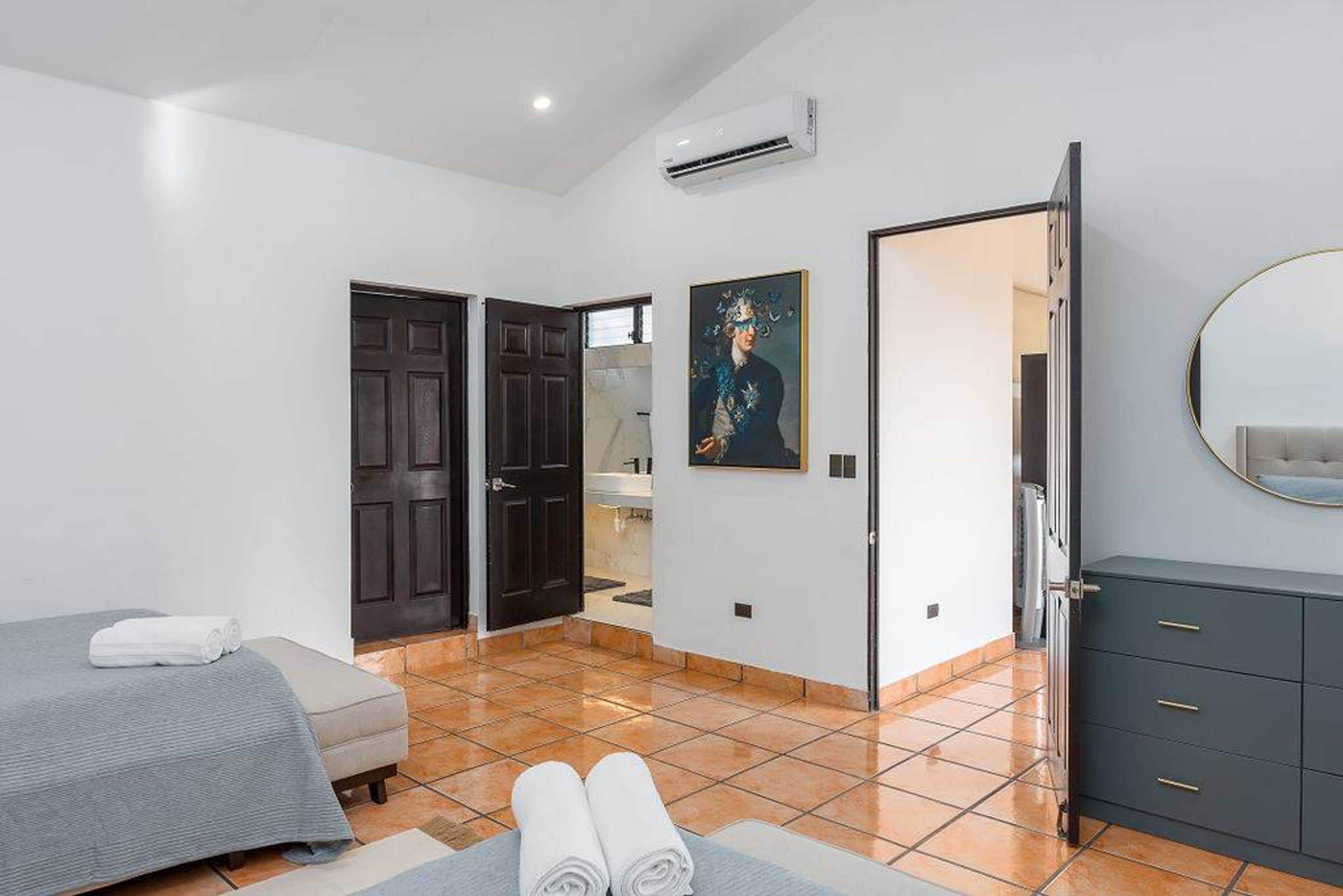 A bedroom with a bed, a dresser, a painting, and a bathroom visible through an open door.