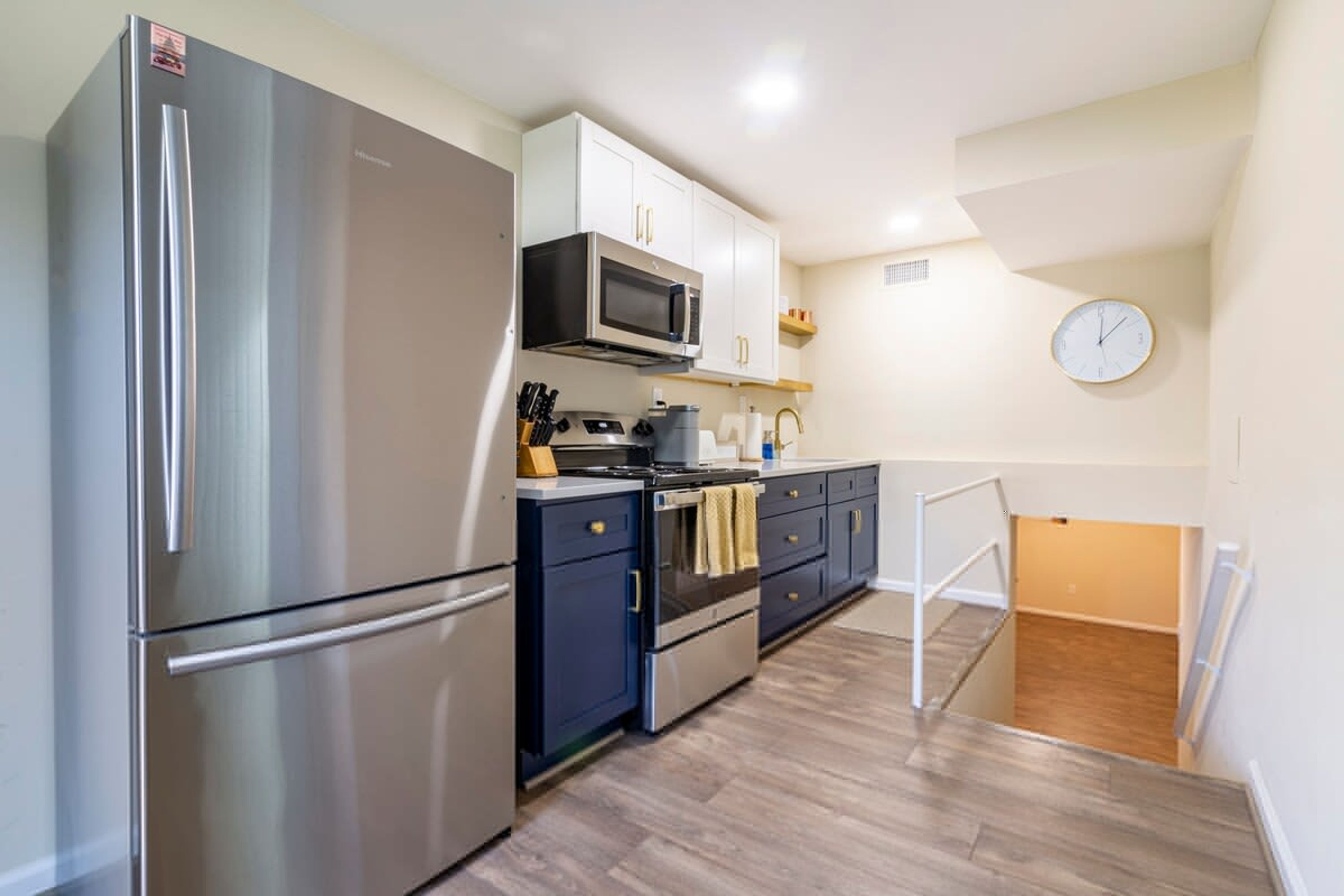 A compact kitchen with a stainless steel refrigerator, a microwave, and a stove. There is a clock on the wall and a small window above the sink.