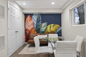 Quarto moderno com sofá de couro marrom, cadeiras brancas, mesa de vidro e parede com padrão de folhas vibrantes.