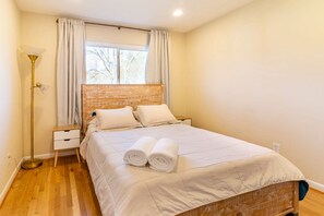 Quarto com cama, mesa de cabeceira, luminária e janela com cortinas.