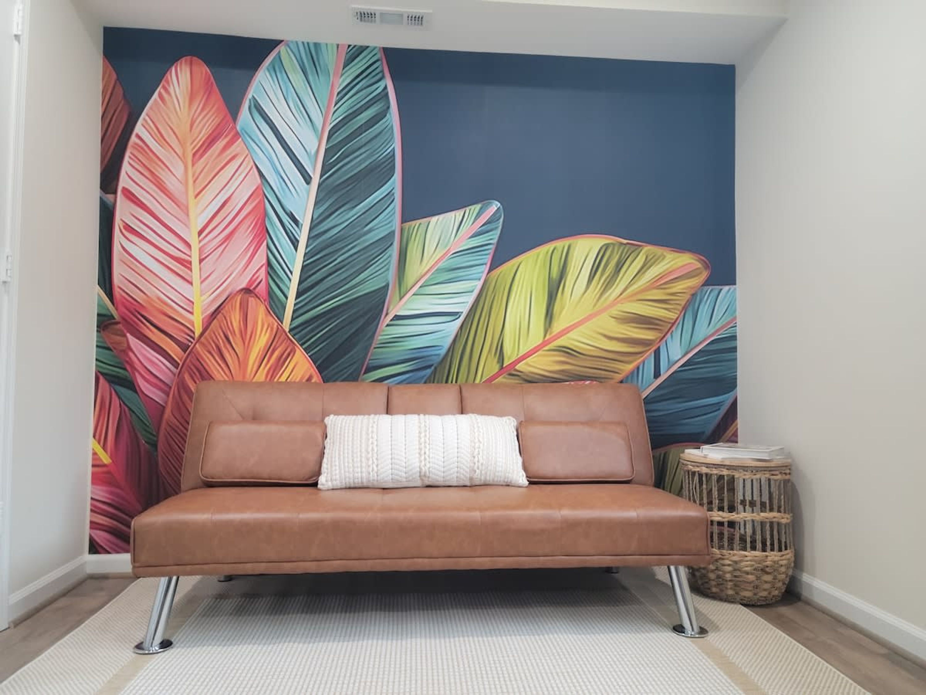 Eine braune Ledersofa mit einem weißen Kissen vor einer Wand mit einem tropischen Blatt-Wandgemälde.