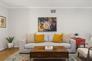 Uma sala de estar com um sofá, duas almofadas amarelas, uma mesa de centro e uma mesa lateral vermelha, além de um vaso com planta.