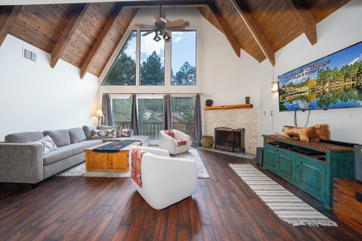 A spacious living room with a high ceiling, wooden beams, a fireplace, a large window, and a television displaying a scenic view.