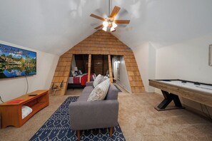 Ein Zimmer mit schrägem Dach, einem Deckenventilator, einem Billardtisch, einem grauen Sofa, einer Holzbank und einem Landschaftsbild.