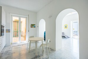 A bright, open-plan living area with a dining table and chairs, a kitchen with a stove, and a view through an archway to a balcony. - Petraio Iconic Terrace on the gulf (Naples)