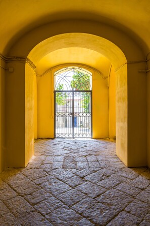 Uma porta arqueada que dá para um pátio com portão de metal, calçamento de pedra e paredes amarelas.