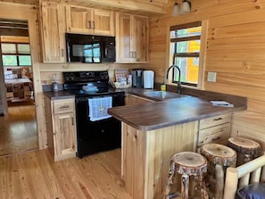 A wooden cabin interior with a kitchen featuring a microwave, stove, and sink. There are stools and a dining area visible.