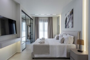 A modern hotel room with a large bed, a flat-screen TV, a bedside lamp, and a view of the city through the window. - Noema Luxury Villas - Villa Calypso, by Estia (Kato Gouves)