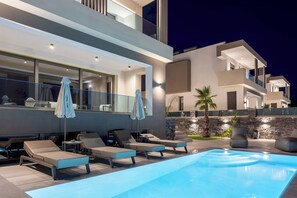Ein moderner Poolbereich im Freien mit Liegestühlen und Sonnenschirmen, umgeben von einem mehrstöckigen Gebäude mit großen Fenstern und einer Steinmauer.