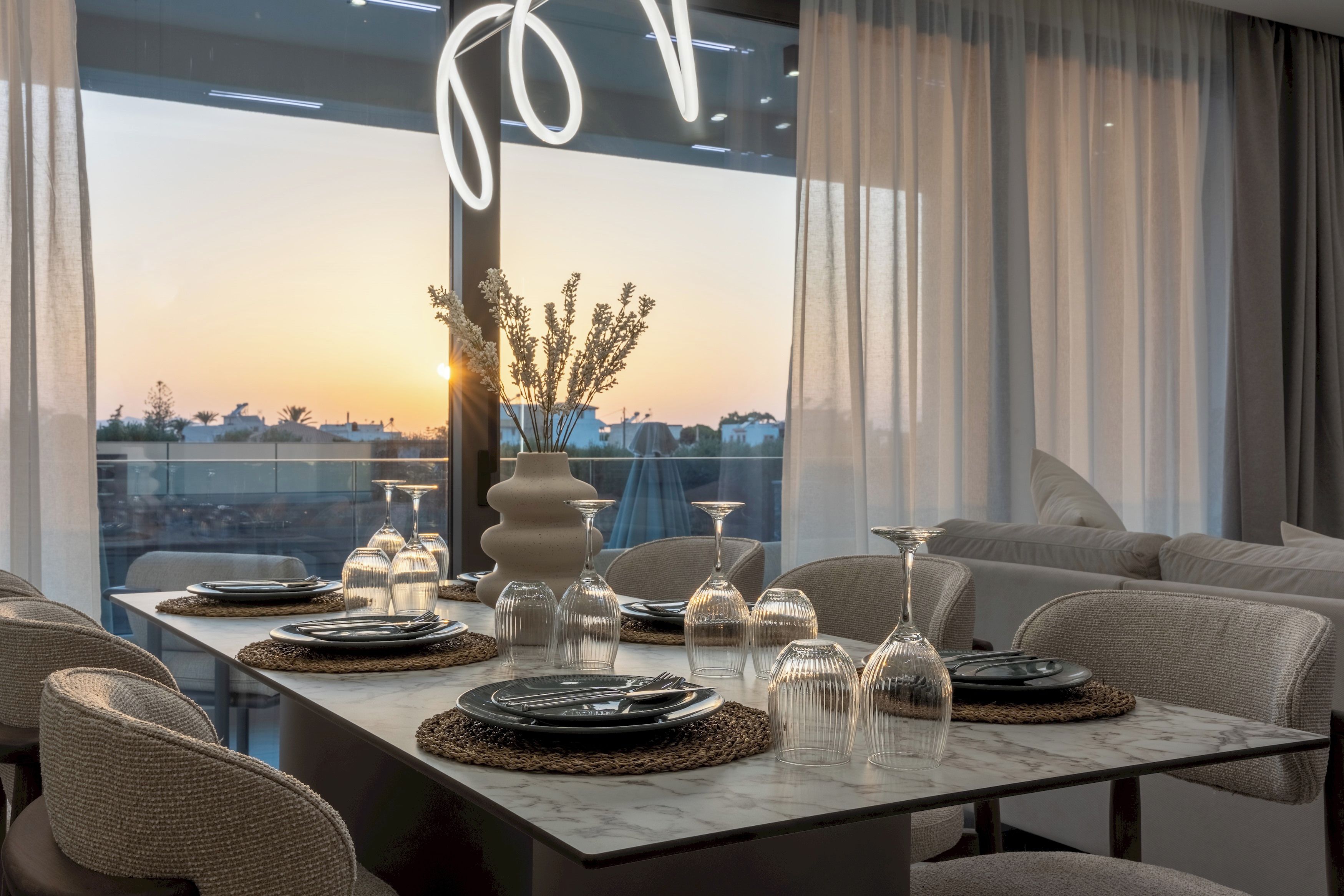 A modern dining area with a table set for four, featuring clear glassware and a vase with dried flowers.