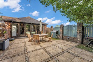 A patio with a brick floor, wooden furniture, and a brick building with large windows. - Oxhay Barn (Newcastle-Under-Lyme)