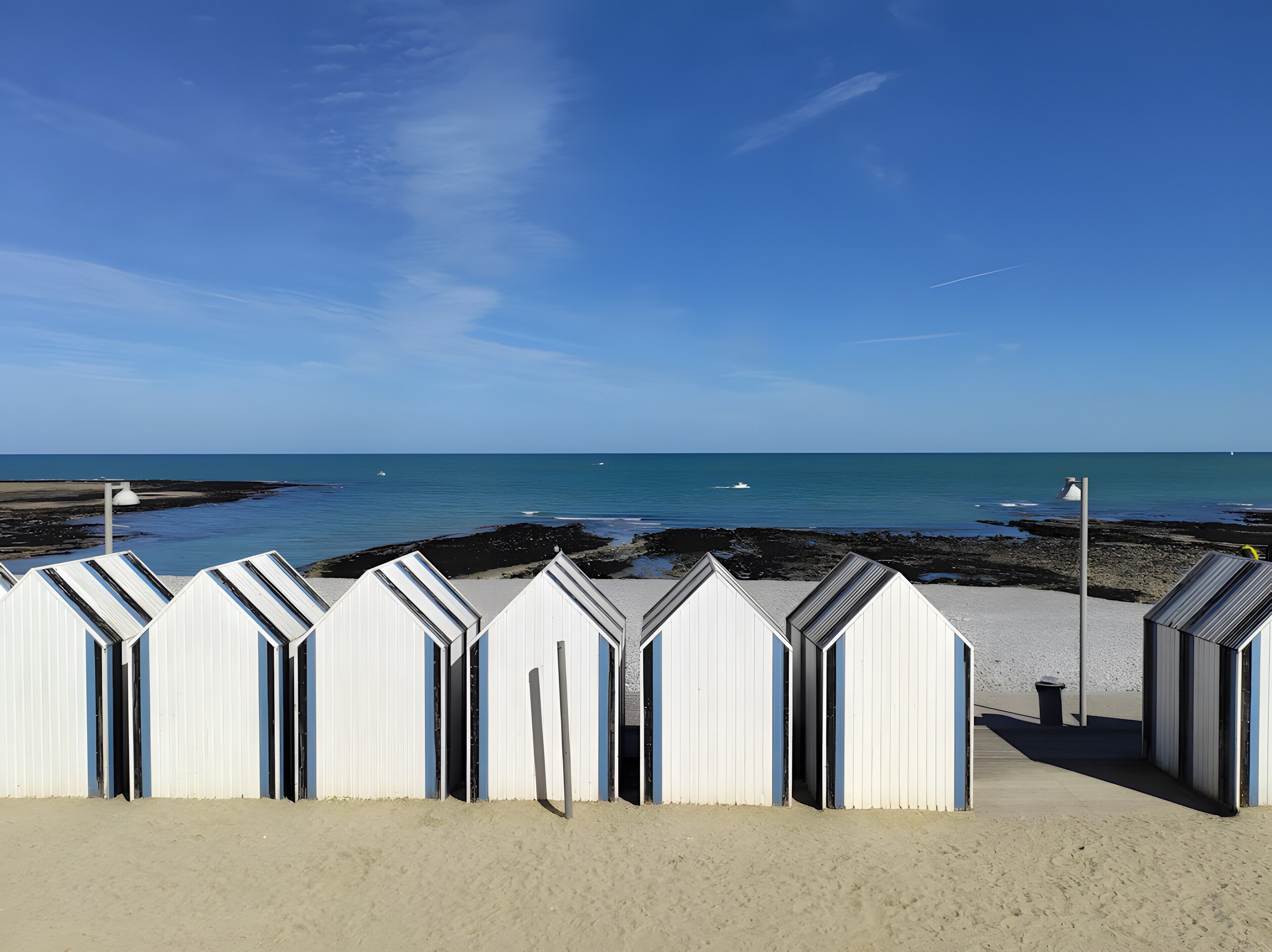 A row of beach huts with open doors, a sandy beach, and a calm sea under a clear sky.