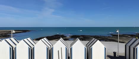 A row of beach huts with open doors, a sandy beach, and a calm sea under a clear sky.