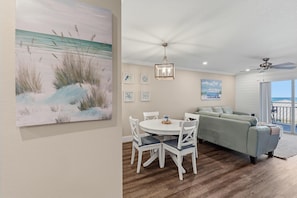 A living room with a dining area, a ceiling fan, and a large painting of a beach scene. - Sea Coast Gardens III 216 (New Smyrna Beach)