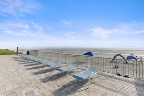 A beachside lounge with sun loungers and umbrellas, a metal railing, and a view of the beach and ocean. - Sea Coast Gardens III 216 (New Smyrna Beach)