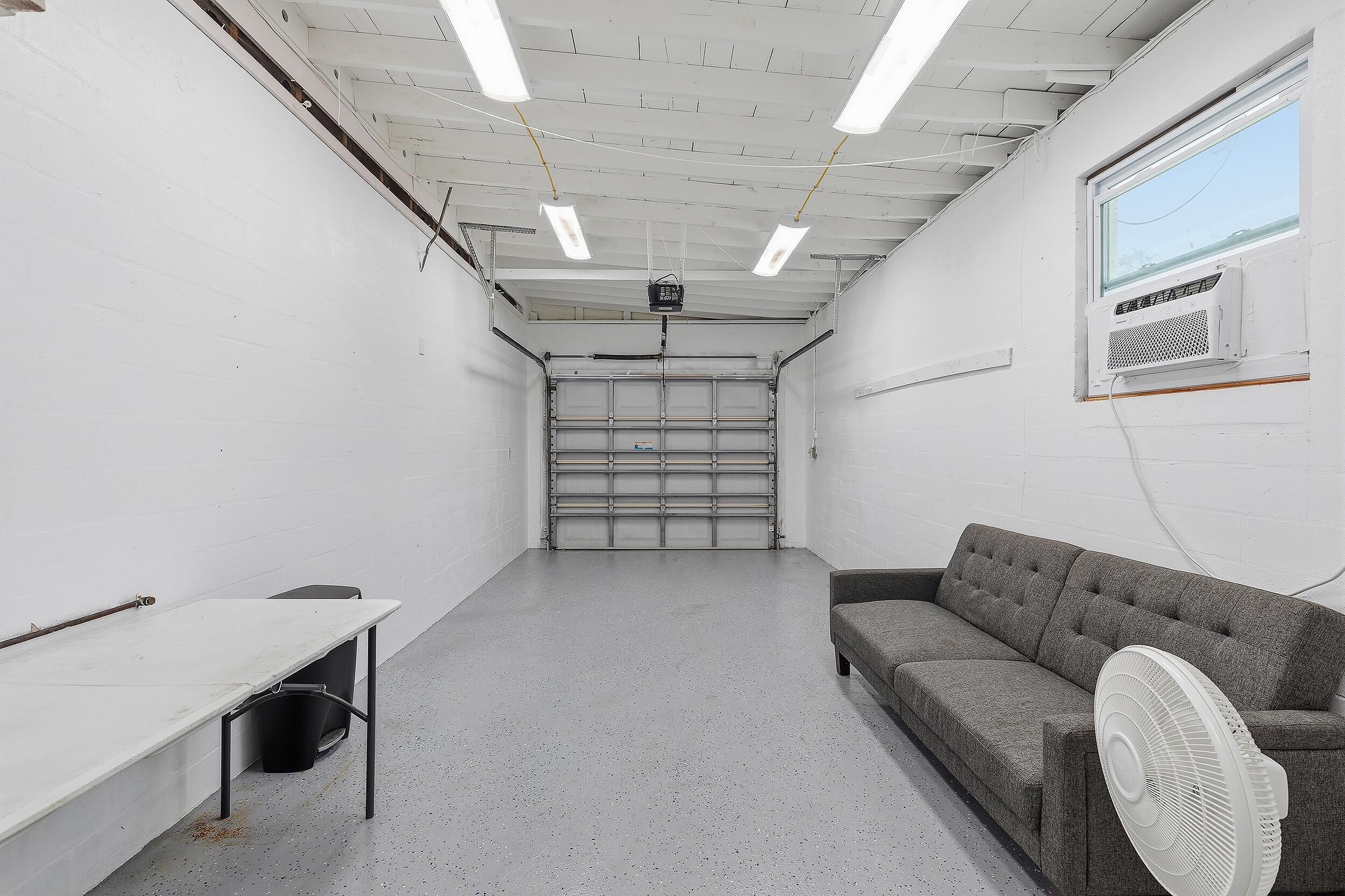 A room with a white ceiling, fluorescent lights, a grey couch, a white fan, and a closed garage door.