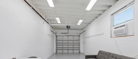 A room with a white ceiling, fluorescent lights, a grey couch, a white fan, and a closed garage door.