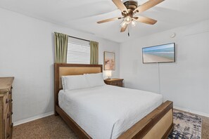 Ein Schlafzimmer mit einem Holzbett, einem Deckenventilator, einem Nachttisch mit Lampe, einem gerahmten Bild und einem an der Wand befestigten Fernseher.