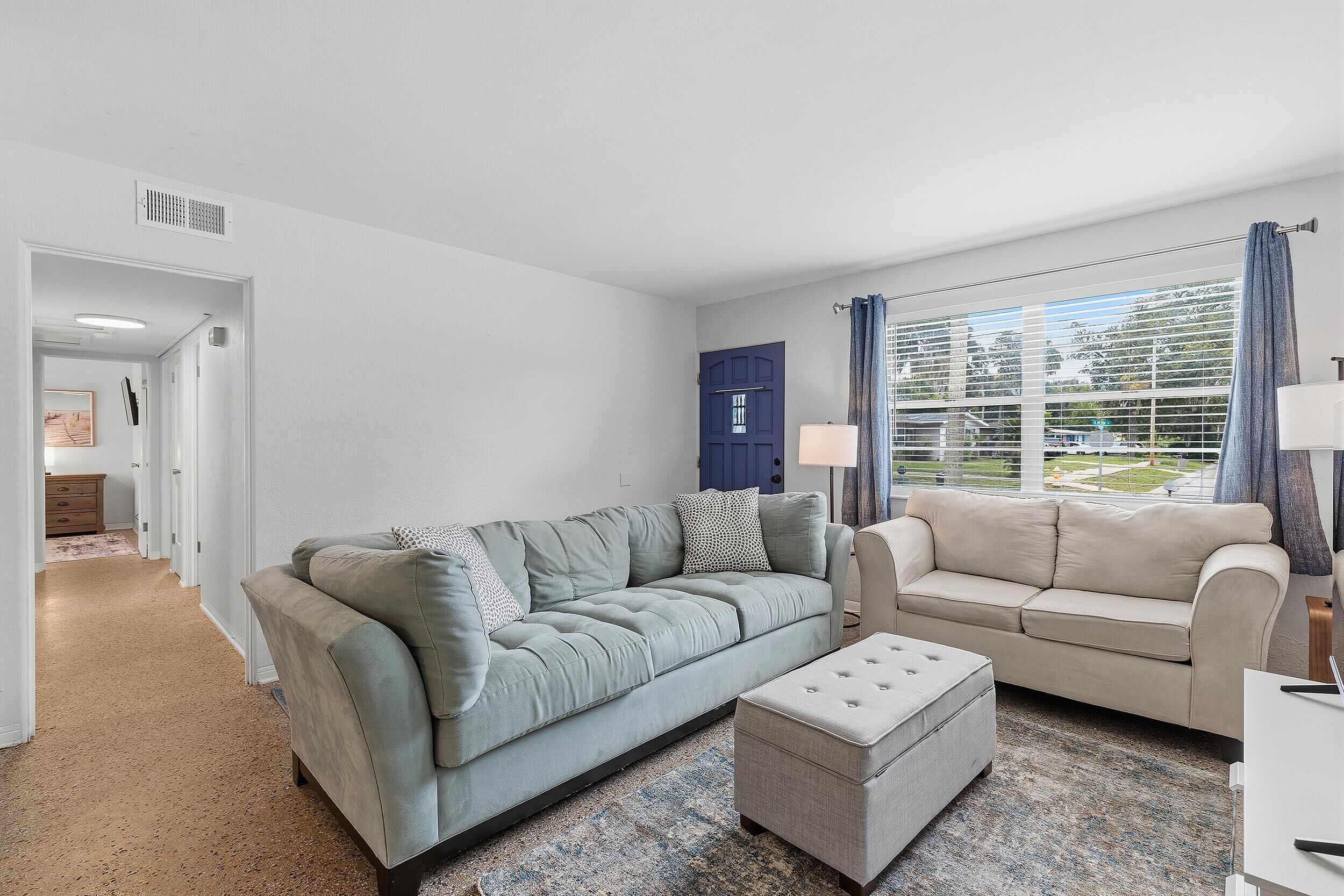 Un salon avec un canapé gris, un pouf assorti, une porte bleue et une fenêtre avec des rideaux.