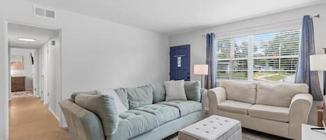 Un salon avec un canapé gris, un pouf assorti, une porte bleue et une fenêtre avec des rideaux.