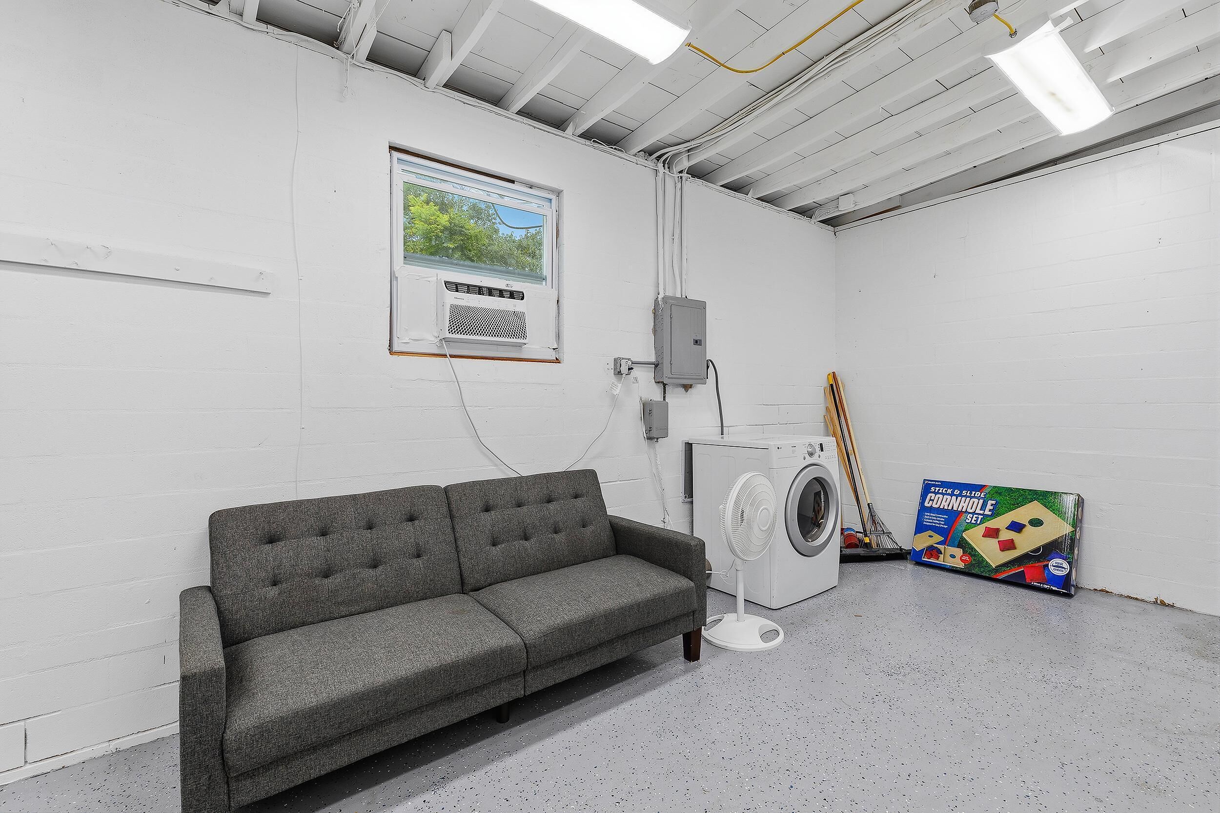 A laundry room with a grey sofa, a washing machine, a fan, and a window with a view of trees.
