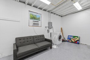 Ein Waschraum mit einer grauen Couch, einer Waschmaschine, einem Ventilator und einem Fenster mit Blick auf Bäume.