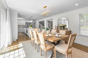 A modern kitchen with white cabinets, a stainless steel refrigerator, and a dining area with a wooden table and chairs. - Retreat Luxury Home West Barnstable (Barnstable)