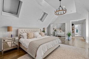A modern bedroom with a large bed, a nightstand, a mirror, and a chandelier. - Retreat Luxury Home West Barnstable (Barnstable)