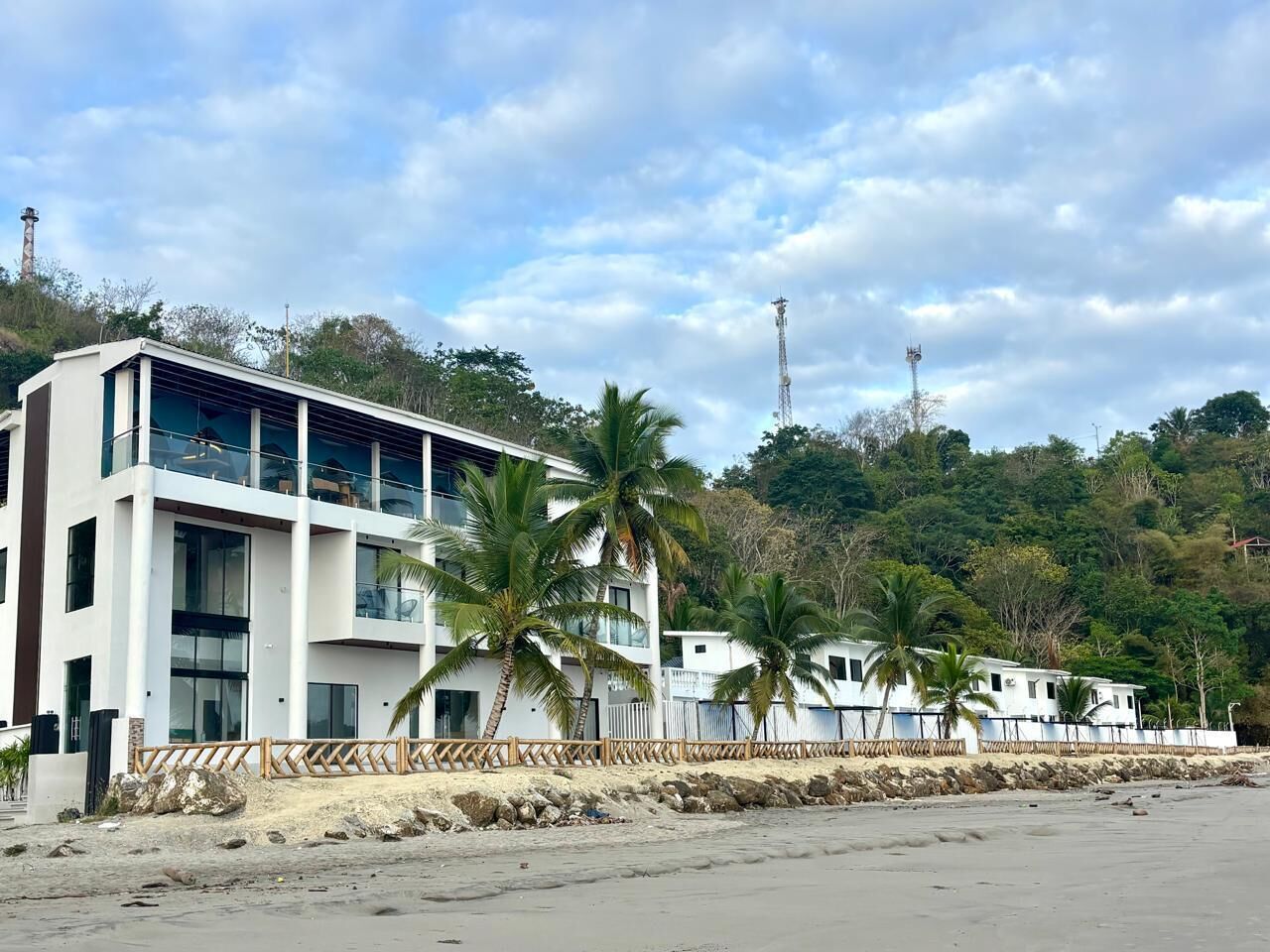 A beachfront hotel with multiple floors, palm trees, and a rocky shoreline.