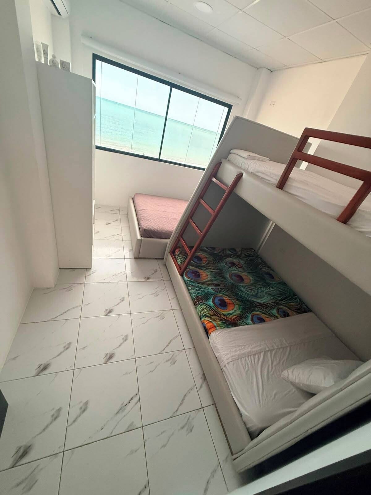A small, well-lit room with a bunk bed, a single bed, a window with a view of the sea, and a tiled floor.