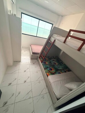 A small, well-lit room with a bunk bed, a single bed, a window with a view of the sea, and a tiled floor. - Hotel Mansion Real (Atacames)