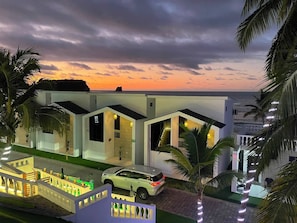 A modern two-story house with a car parked in front, surrounded by palm trees and a view of the ocean at sunset. - Hotel Mansion Real (Atacames)
