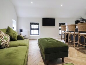 Ein modernes Wohnzimmer mit einem grünen Sofa, passendem Hocker, einer Essecke mit Korbstühlen und einem großen Flachbildfernseher an der Wand.
