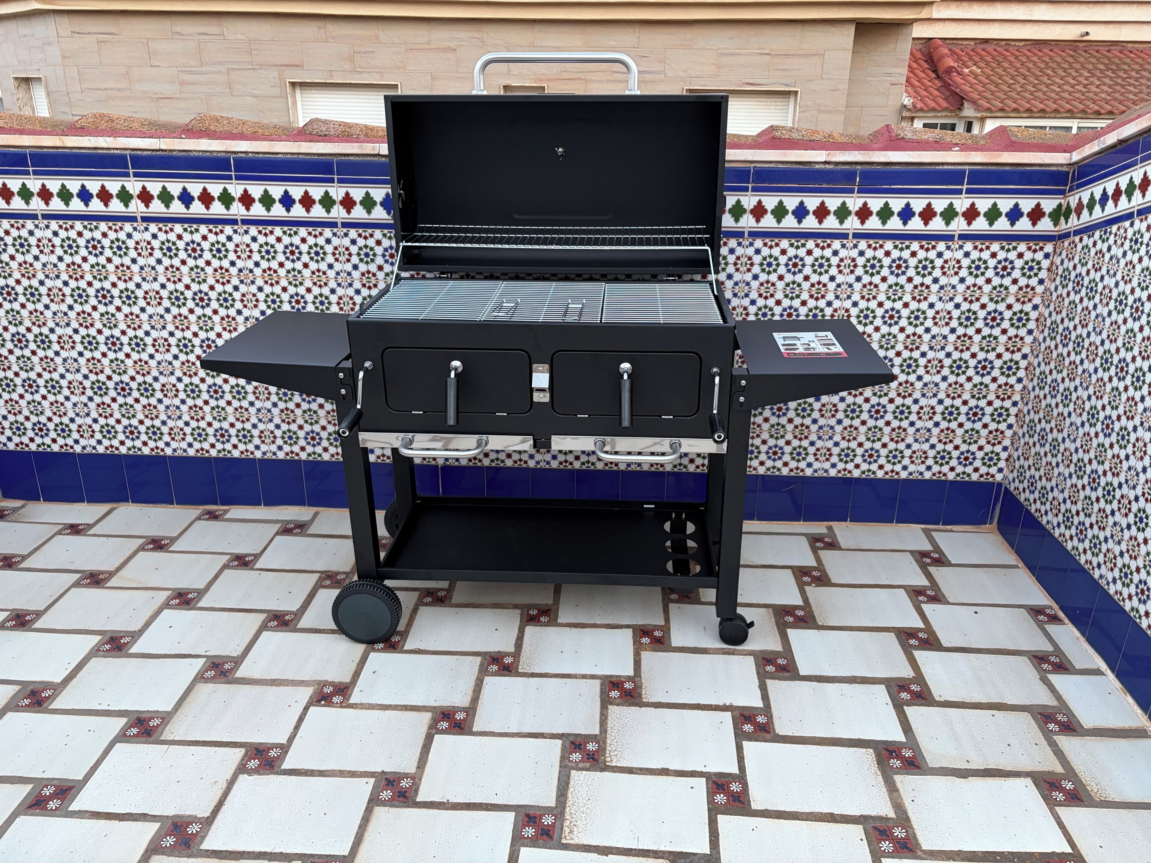 Una parrilla con tapa negra y varios quemadores, ubicada en un patio empedrado con un borde decorativo.