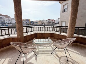 A balcony with two wicker chairs and a glass table, overlooking a cityscape with buildings and a clear sky.